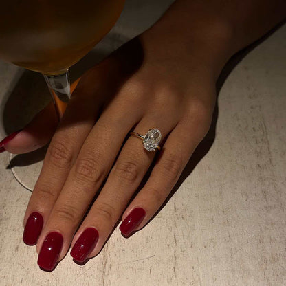 Hand wearing Deus Ring Pherinwen with oval-cut moissanite solitaire and deep red nails holding glass