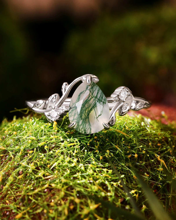 Pear-shaped Moss Agate engagement ring with Moissanite accents and leaf-inspired silver band on moss background