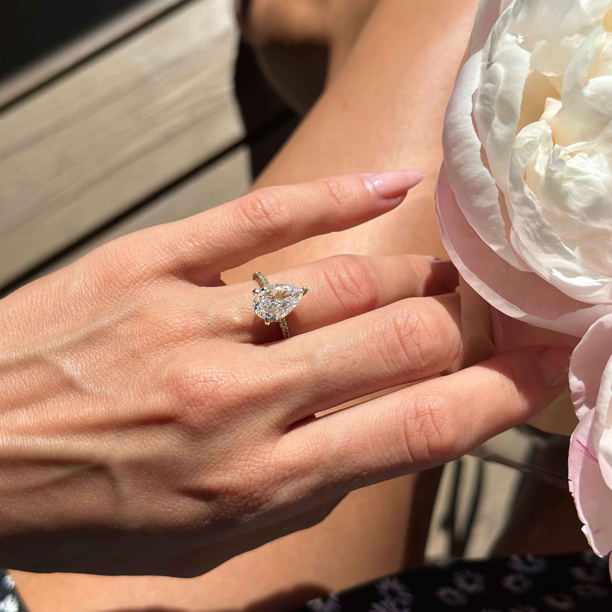 Kylariel solitaire engagement ring with pear-shaped moissanite and half pavé design on hand near white flower