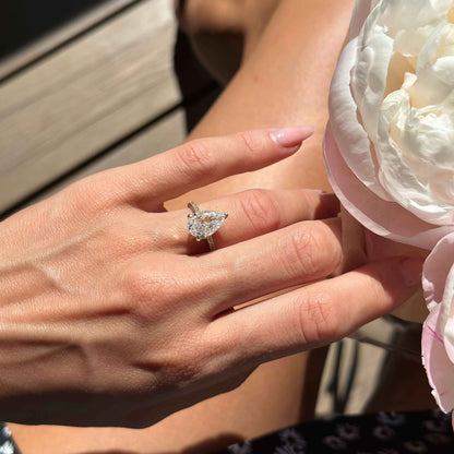 Kylariel solitaire engagement ring with pear-shaped moissanite and half pavé design on hand near white flower